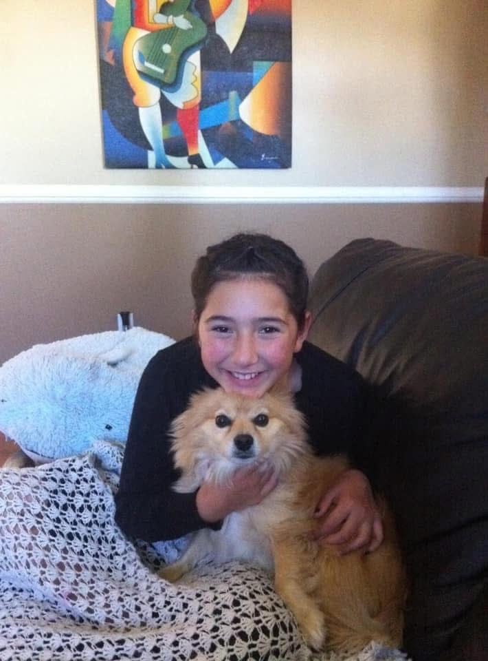 A smiling 10-year-old girl on a couch hugging a small tan dog